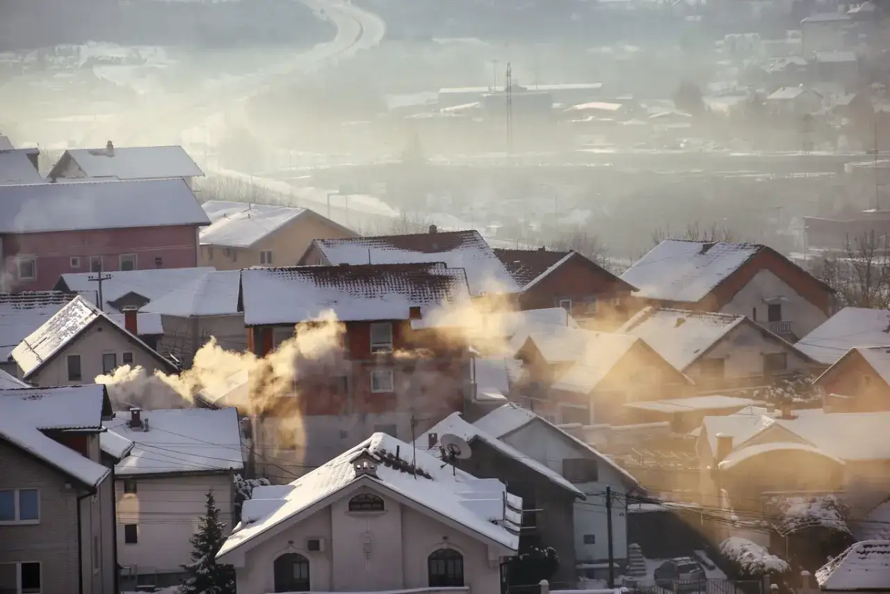 Dym z kominów domów pokrytych śniegiem unosi się w powietrze, pokazując, jak powstaje smog w zimowy poranek.